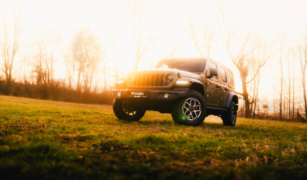 Zielony Jeep Wrangler Rubicon z włączonymi reflektorami zaparkowany na trawie o zachodzie słońca.