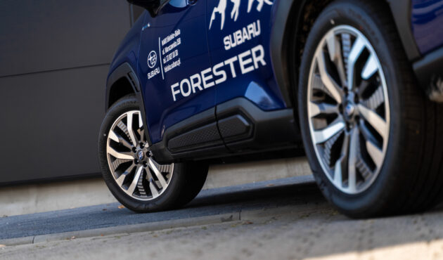 Zbliżenie na przednie koło i felgę alufelgową niebieskiego samochodu Subaru Forester z widocznym napisem modelowym i grafiką gór.